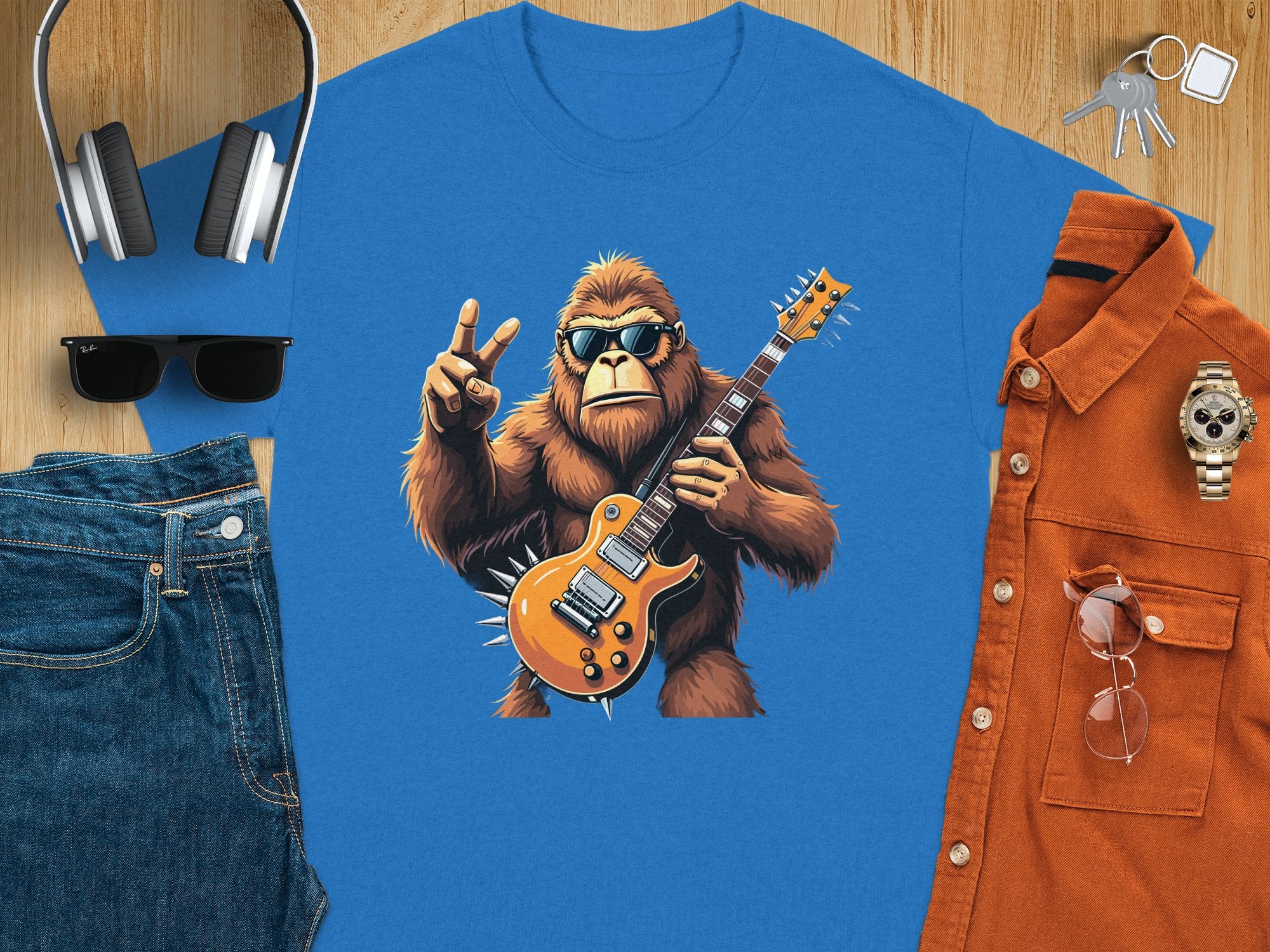 Garment Graphics offers a blue t-shirt for music lovers featuring a gorilla playing an electric guitar and flashing a peace sign. Its styled with jeans, sunglasses, headphones, keys, an orange button-up shirt, and a wristwatch on a wooden surface.