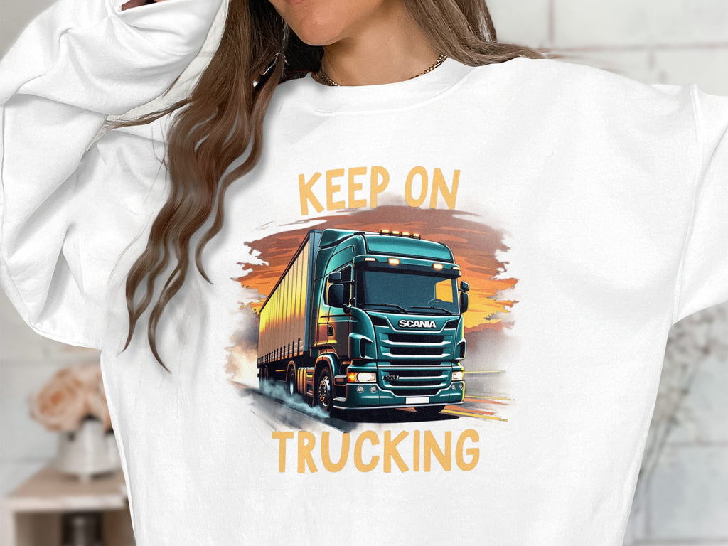 A truck enthusiast wears a Garment Graphics white sweatshirt featuring a green truck under a sunset, emblazoned with KEEP ON TRUCKING. It embodies adventure and freedom for open road lovers.