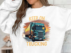 A truck enthusiast wears a Garment Graphics white sweatshirt featuring a green truck under a sunset, emblazoned with KEEP ON TRUCKING. It embodies adventure and freedom for open road lovers.