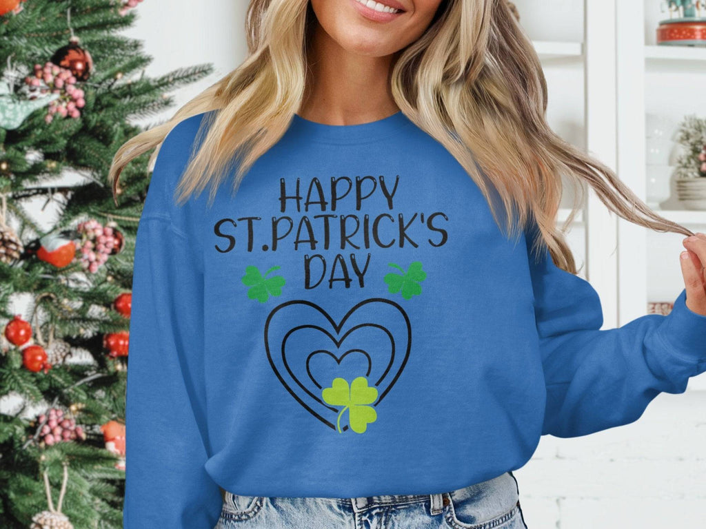 Someone is wearing a blue festive sweatshirt from Garment Graphics with Happy St. Patricks Day printed in black, highlighted by green clover designs and hearts, with a decorated Christmas tree as the backdrop.