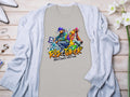 A vibrant light gray t-shirt from Garment Graphics features a street dance graphic of two dancers and the text Risk Taker above Street Dance with Fran, celebrating the Risk Taker 10th Final T-Shirts. Displayed on wood, it pairs with a blue cardigan, accessories, and white flowers.