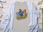 A vibrant light gray t-shirt from Garment Graphics features a street dance graphic of two dancers and the text Risk Taker above Street Dance with Fran, celebrating the Risk Taker 10th Final T-Shirts. Displayed on wood, it pairs with a blue cardigan, accessories, and white flowers.