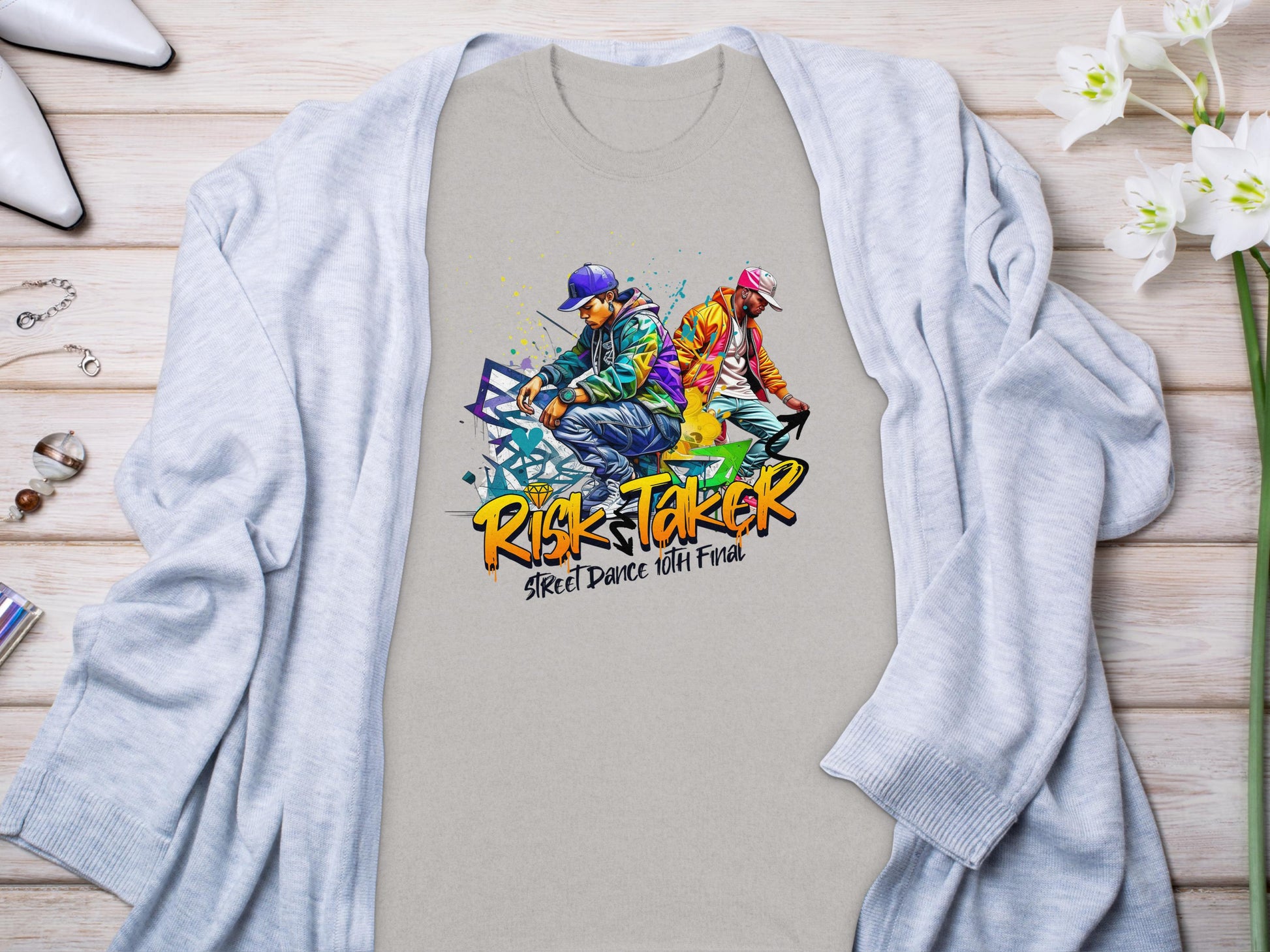 A vibrant light gray t-shirt from Garment Graphics features a street dance graphic of two dancers and the text Risk Taker above Street Dance with Fran, celebrating the Risk Taker 10th Final T-Shirts. Displayed on wood, it pairs with a blue cardigan, accessories, and white flowers.