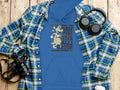 A Garment Graphics blue classic fit hoodie, featuring a gator graphic and See You Later Alligator, is beneath a blue and green plaid shirt. A camera, headphones, and white coffee cup are nearby on the wooden surface.