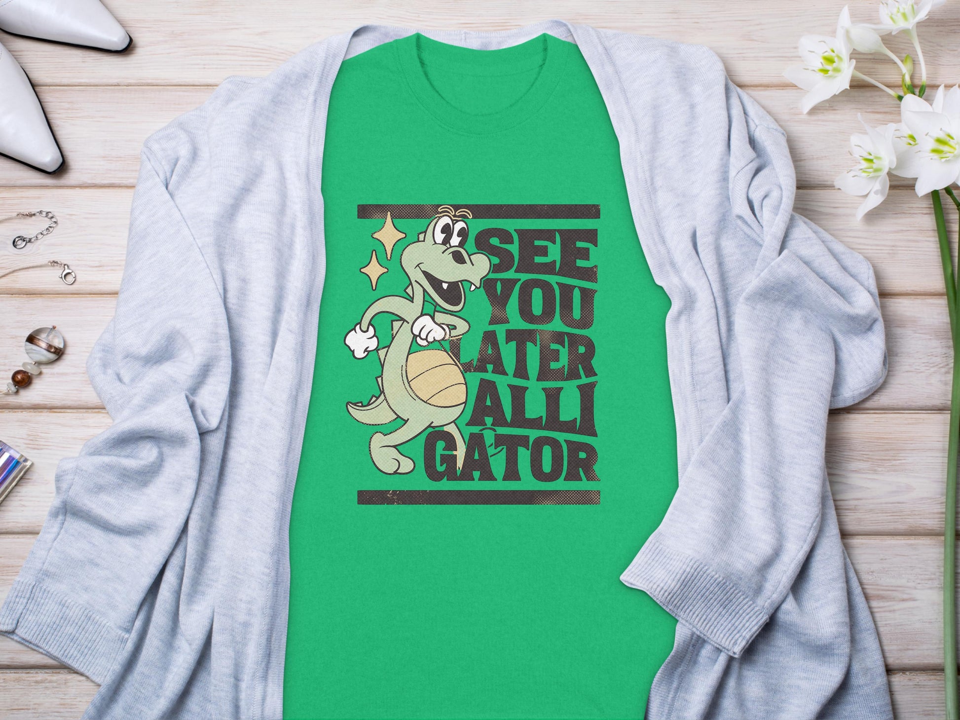 A Garment Graphics green T-shirt showcases a cartoon alligator with a basketball and See You Later Alligator text. Ideal for casual wear, its on a wooden surface, partially covered by a light gray cardigan, with shoes and flowers adding to the relaxed vibe.