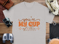 A white 100% cotton Garment Graphics T-shirt, featuring Youre My Cup of Tea in orange cursive, is placed on a wooden surface. Nearby are a brown camera case, vintage camera, binoculars, belt, and canvas bag.