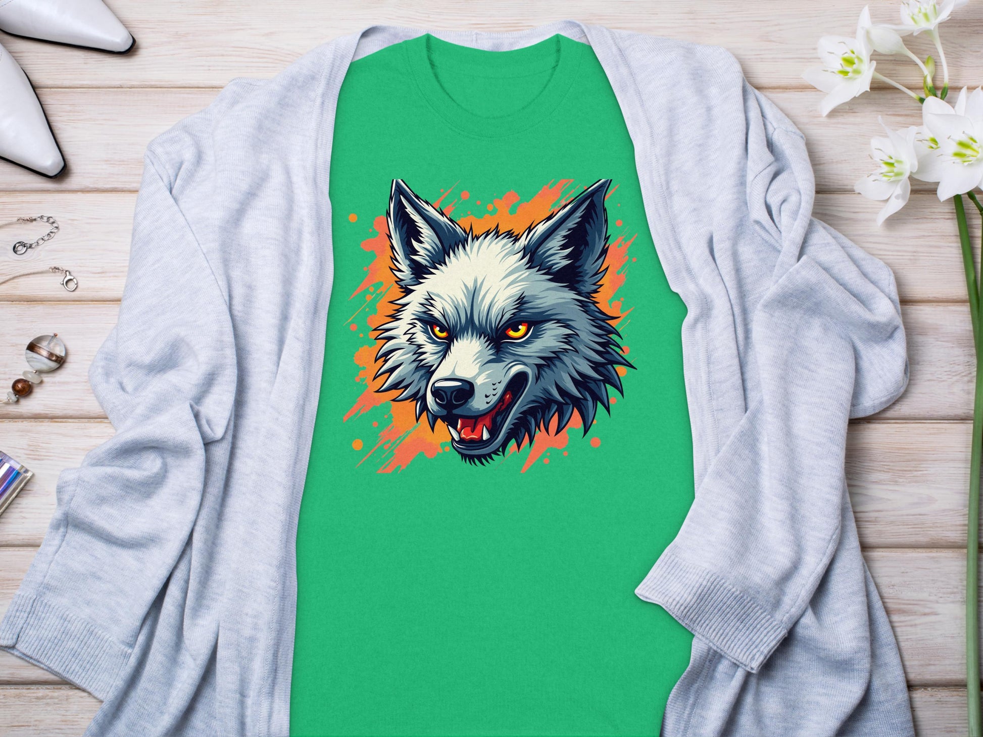 Garment Graphics green t-shirt showcases a fearless wolf graphic with intense eyes. Its neatly placed on a light wooden surface, partially covered by a gray cardigan, surrounded by white flowers, stylish white shoes, and delicate jewelry items.