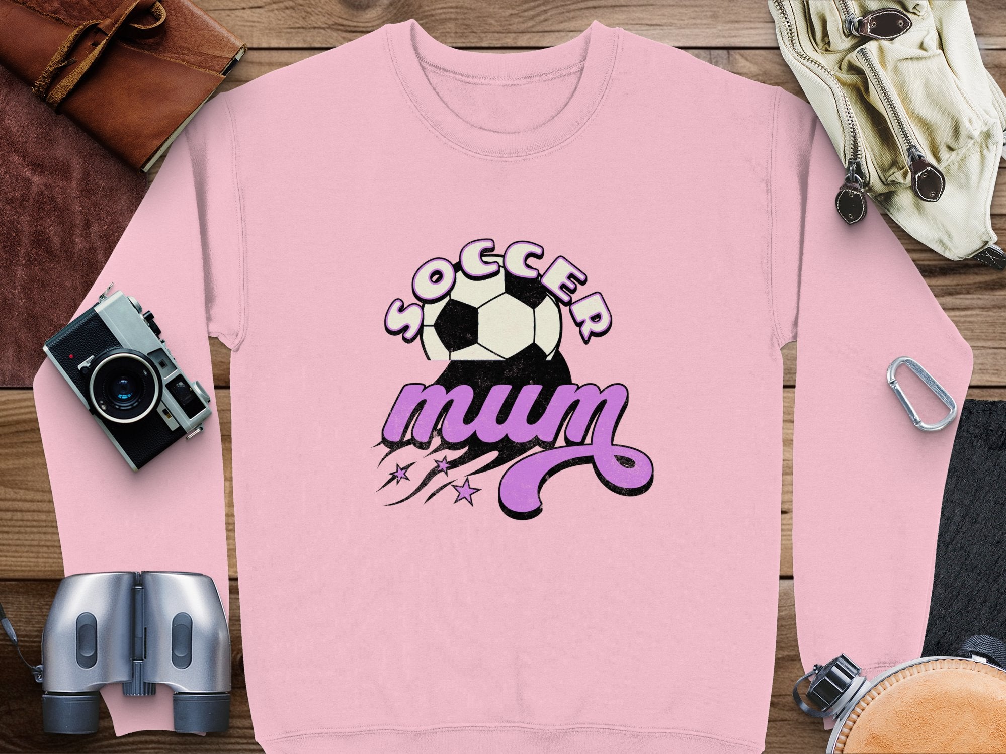 A Garment Graphics pink sweatshirt with a vintage soccer ball design and Soccer Mum text lies flat on wood, surrounded by binoculars, a camera, a belt, and a bag, creating an appealing sporty scene.