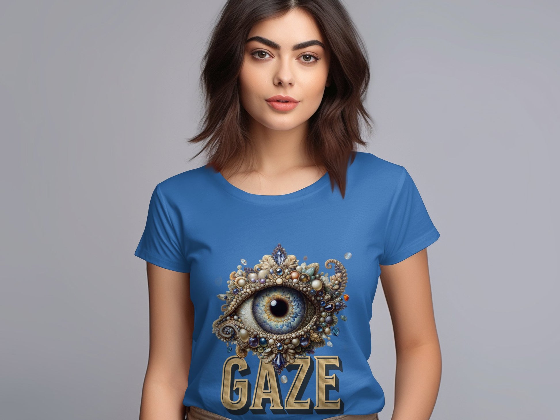 A woman with shoulder-length brown hair wears a blue Garment Graphics T-shirt featuring a large eye adorned with jewels and details. GAZE is elegantly inscribed below. She stands against a gray background, showcasing it in womens sizing effortlessly.
