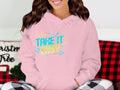 A person wearing a classic fit pink hoodie by Garment Graphics featuring TAKE IT EASY in bold letters, colorful arrows, and musical notes for a comfy fashion vibe. The background includes a red and black checkered pattern and part of a Christmas Tree pillow.