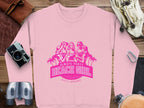 Garment Graphics introduces a pink sweatshirt adorned with Always Party Beach Girl text and illustrations of three women amid palm trees. Nearby, on a wooden surface, lie items such as a camera, binoculars, bag, scarf, and carabiner.