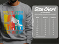A person wears a gray Garment Graphics DJ sweatshirt with a vibrant graphic reading Dont Stop the Music, ideal for music lovers. Next to them is a Gildan 18000 heavy blend crewneck unisex size chart with sizes and measurements in inches.