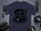 Celebrate in style with Garment Graphics navy blue T-shirt adorned with a black top hat, two shamrocks, and St Patricks Day text. A smartphone, smartwatch, shoes, dumbbells, and a portable speaker create a festive scene against a gray background.