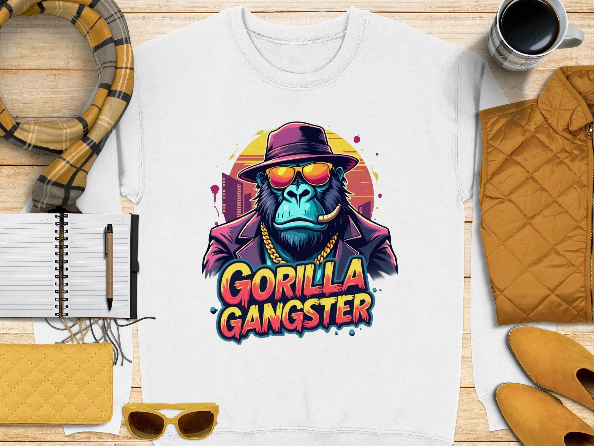 A Garment Graphics white sweatshirt captures Urban Style with a vivid graphic of a gorilla in sunglasses, hat, and chain necklace labeled Gorilla Gangster, showcased on wood with a scarf, notebook, pen, yellow purse, and coffee cup for added chicness.