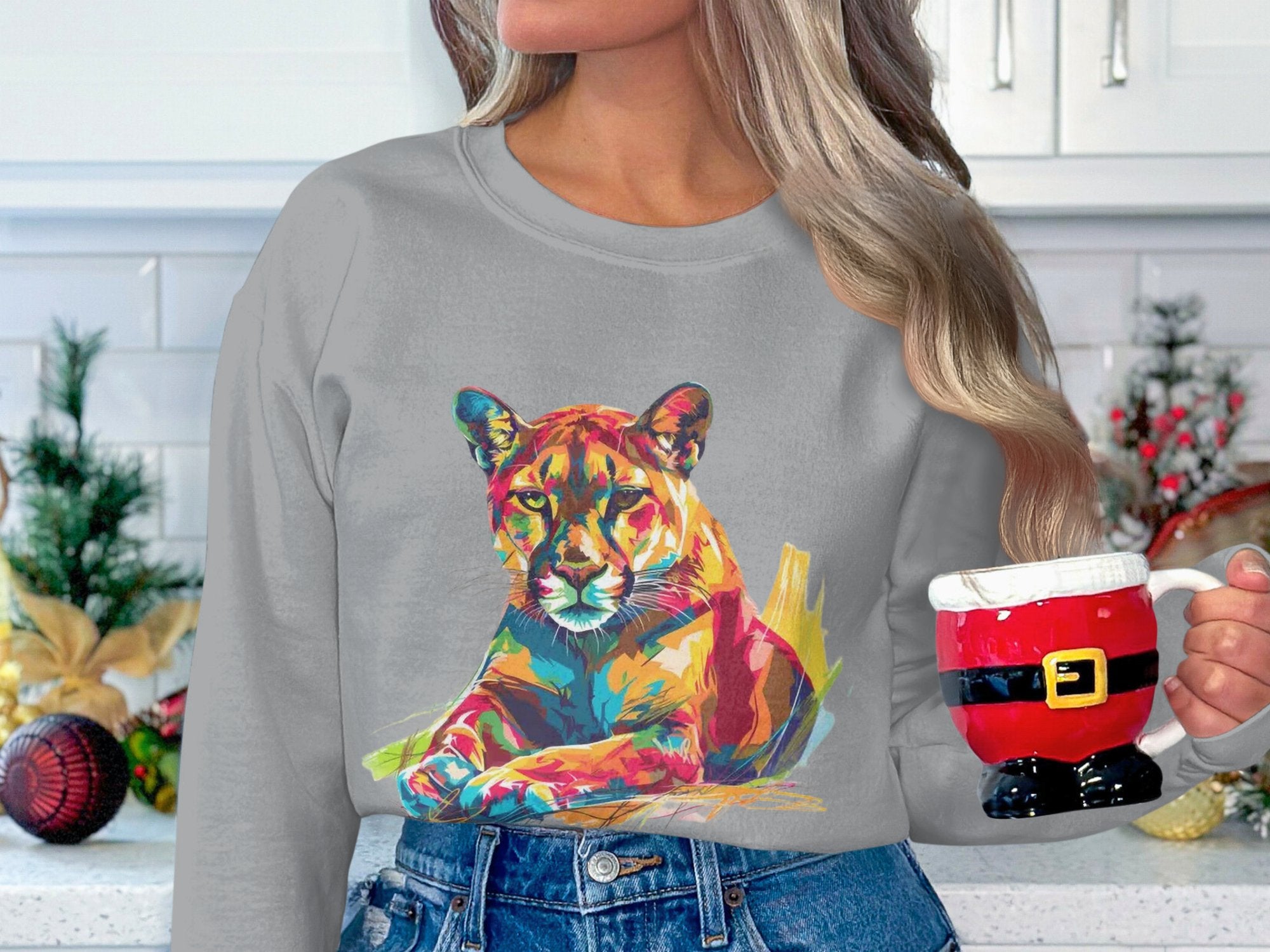 In a festive kitchen, a person with long hair holds a Santa-themed mug. They wear a gray sweater featuring a vibrant big cat design by Garment Graphics, which complements the holiday decorations against the white tiled wall.