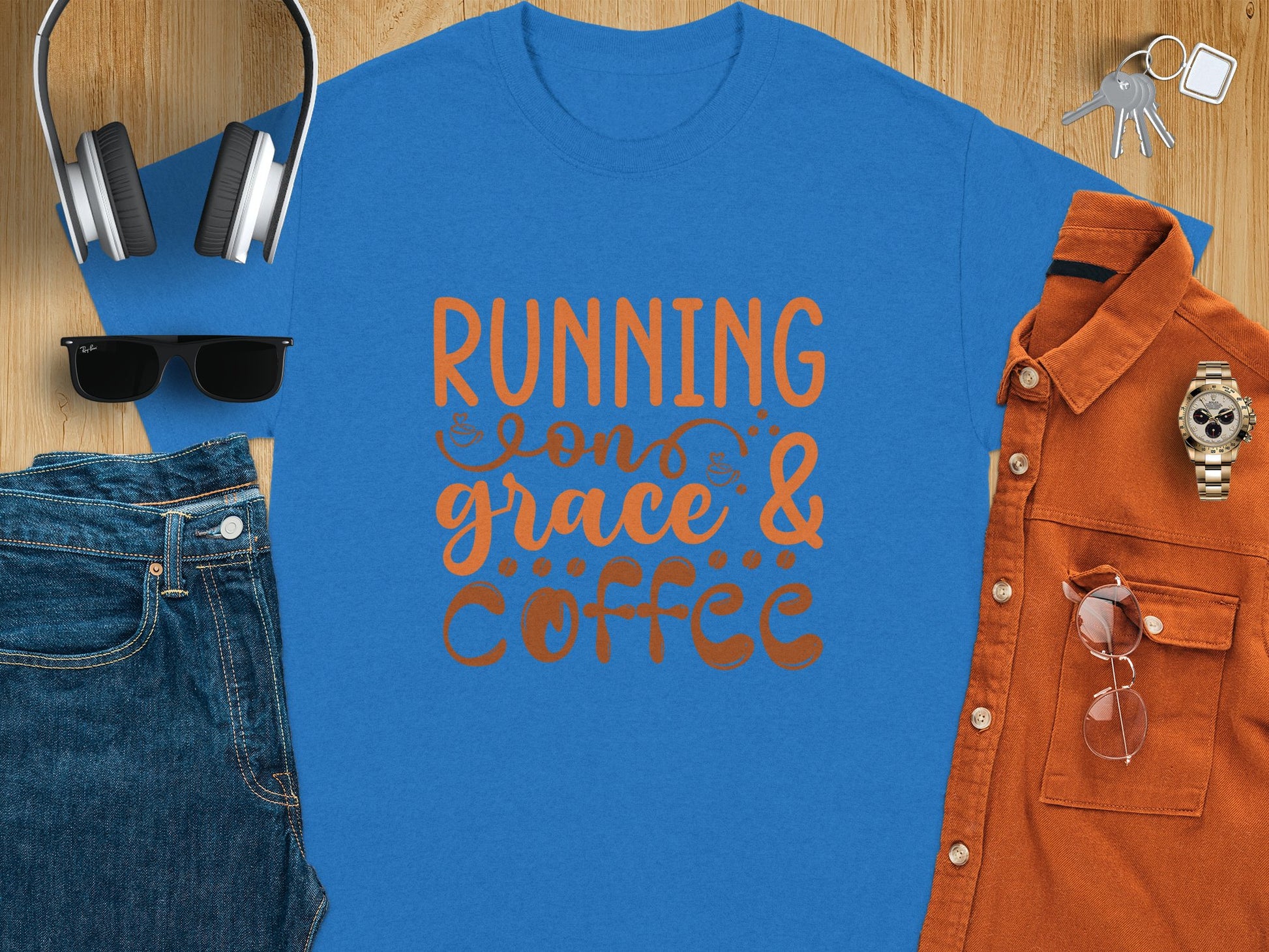 Flat lay featuring Garment Graphics blue graphic t-shirt with Running on Grace & Coffee in bold orange. Accompanied by essentials like headphones, keys, a wristwatch, sunglasses, an orange shirt, and blue jeans, all gracefully arranged on a wooden surface.