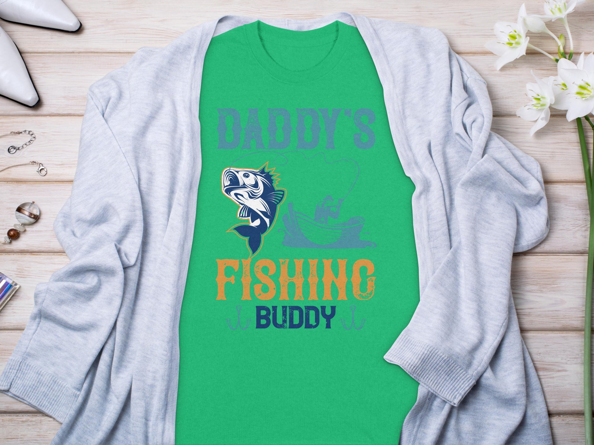 A green Graphic Print T-Shirt from Garment Graphics, featuring Daddys Fishing Buddy with a fish and person in a boat. Styled on wood with a gray cardigan, it’s perfect for fishing fans and is surrounded by flowers, jewelry, and white heels.