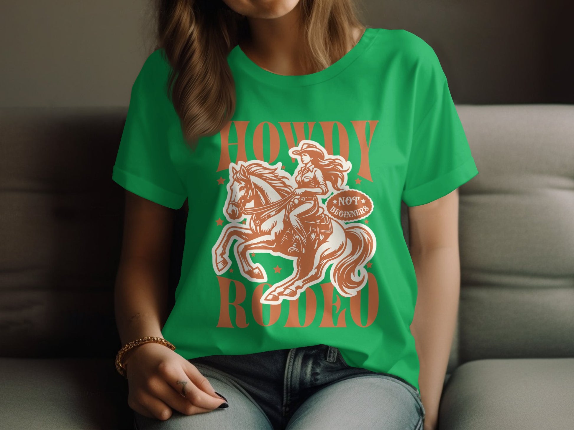 A person wearing a green Garment Graphics Rodeo T-Shirt with a cowboy illustration and Howdy Rodeo text sits on the couch, hands resting casually in their lap. The shirt is available in womens sizing.