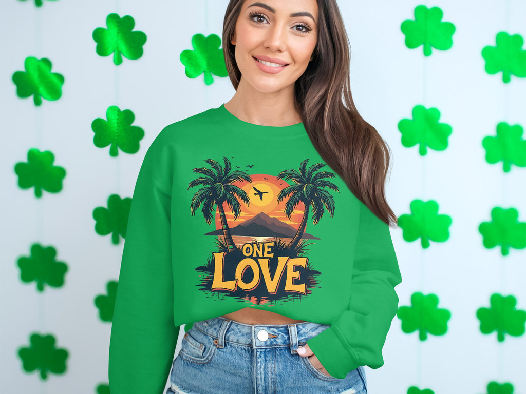 A person in a green Garment Graphics sweatshirt featuring the One Love Sunset graphic with vibrant palm trees stands before a shamrock wall. With long hair flowing and denim shorts completing their outfit, they capture a sunny, carefree vibe.