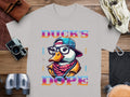 The Garment Graphics Ducks Dope T-Shirt displays a vibrant duck illustration with glasses, cap, and scarf. Bold gradient Ducks Dope text stands out. Its set on wood with cameras, binoculars, and a bag nearby, showcasing its unique style.