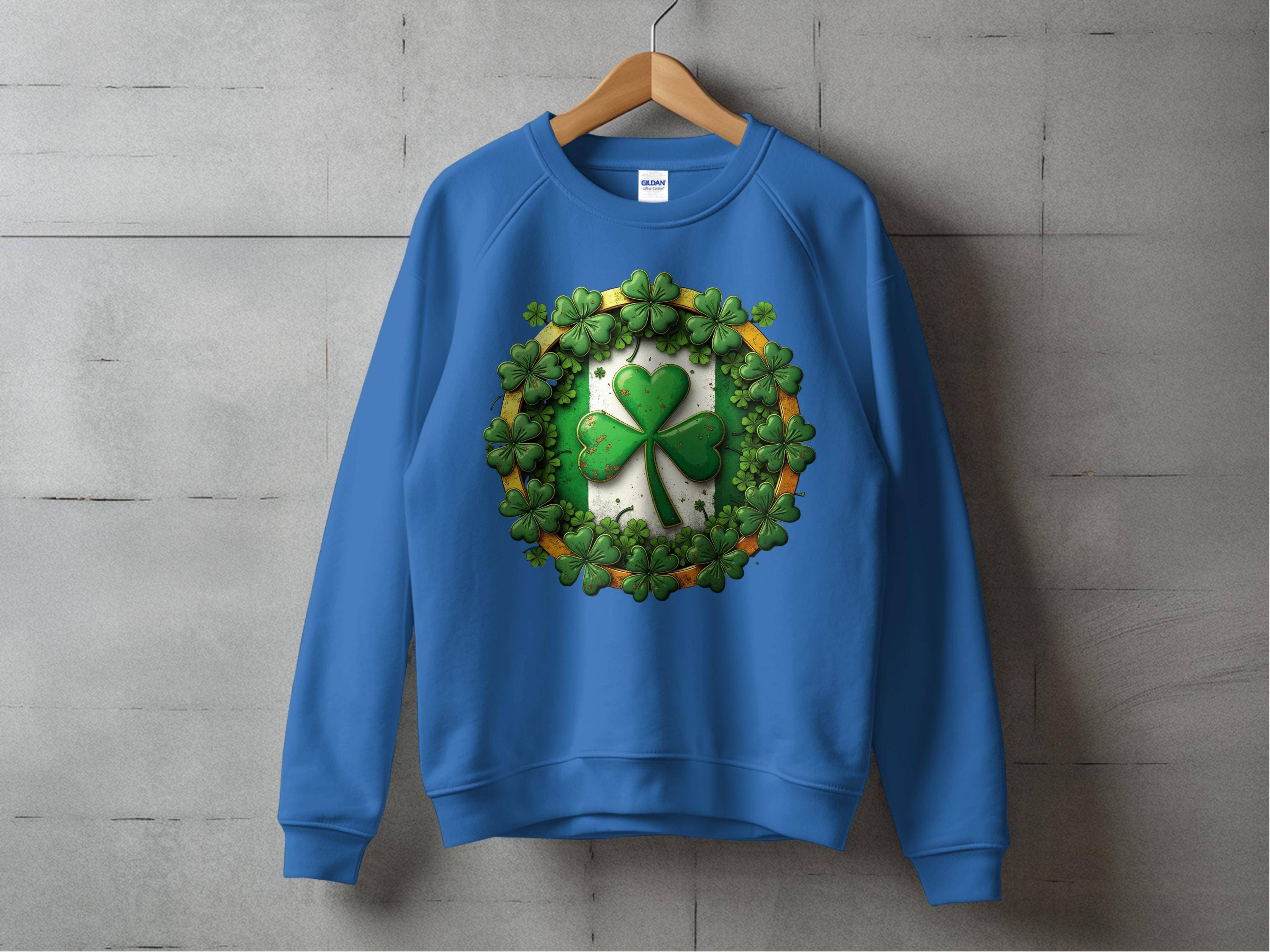 A Garment Graphics blue sweatshirt, embodying Irish spirit, hangs on a wooden hanger against a concrete wall. It features a circle design with a green shamrock at the center, surrounded by smaller shamrocks, encapsulating luck and tradition.