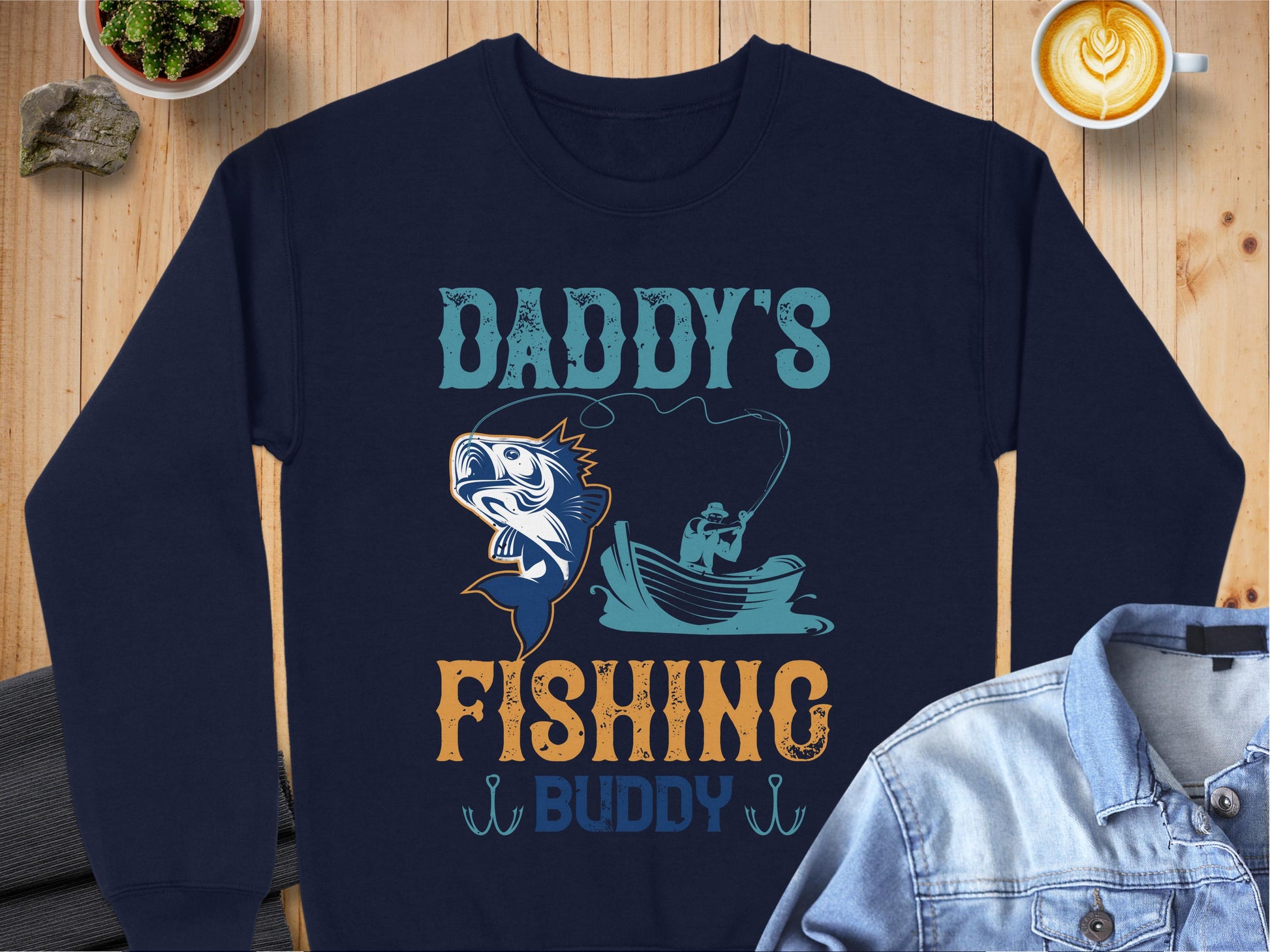 A navy blue sweatshirt by Garment Graphics, ideal for fishing fans, lies on a wooden surface. It boasts the Daddys Fishing Buddy design with a boat fishing scene and large fish illustration. Nearby are a denim jacket, latte, and small plant.