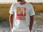 A person wears the Born To Be Musician T-shirt from Garment Graphics, ideal for music enthusiasts. It showcases bold lettering with vibrant drummer and guitarist silhouettes in orange and red on grassy dunes.