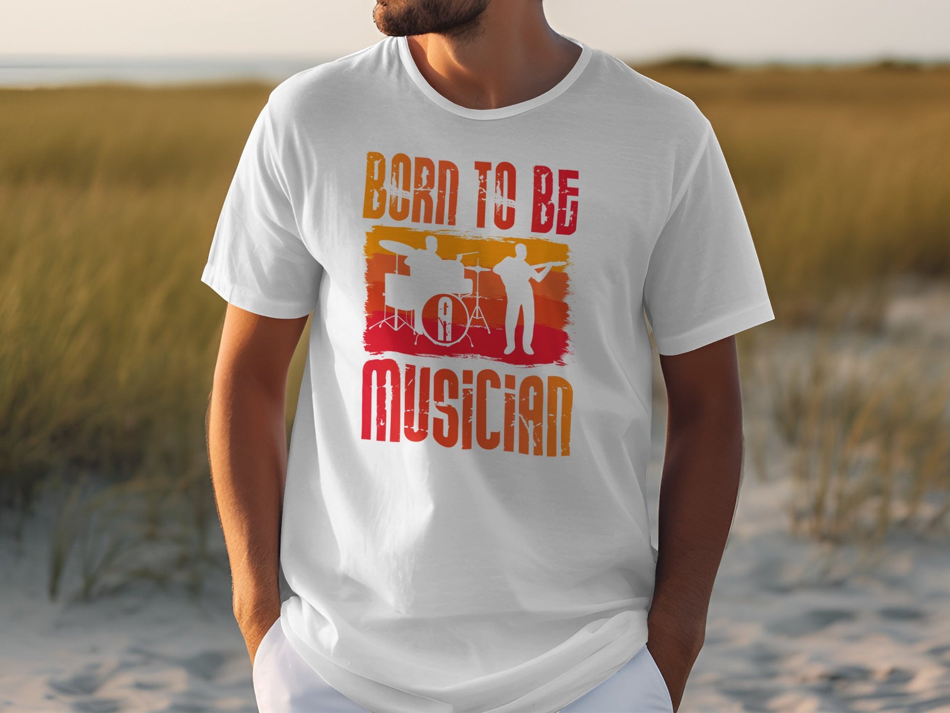 A person wears the Born To Be Musician T-shirt from Garment Graphics, ideal for music enthusiasts. It showcases bold lettering with vibrant drummer and guitarist silhouettes in orange and red on grassy dunes.