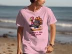 A man on the beach wears a pink T-shirt from Garment Graphics, featuring a whimsical cartoon turkey and the text Turkey on the Run. The ocean and sand form a serene backdrop for flaunting the humorous graphic design.