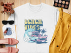 Garment Graphics offers a retro white sweatshirt titled Beach Vibes with a classic car, waves, palm trees, and sunset design. Styled on wooden floor; paired with a mustard yellow blazer, pink shoes, makeup brushes, eyeshadow palette, sunglasses, and nail polish.