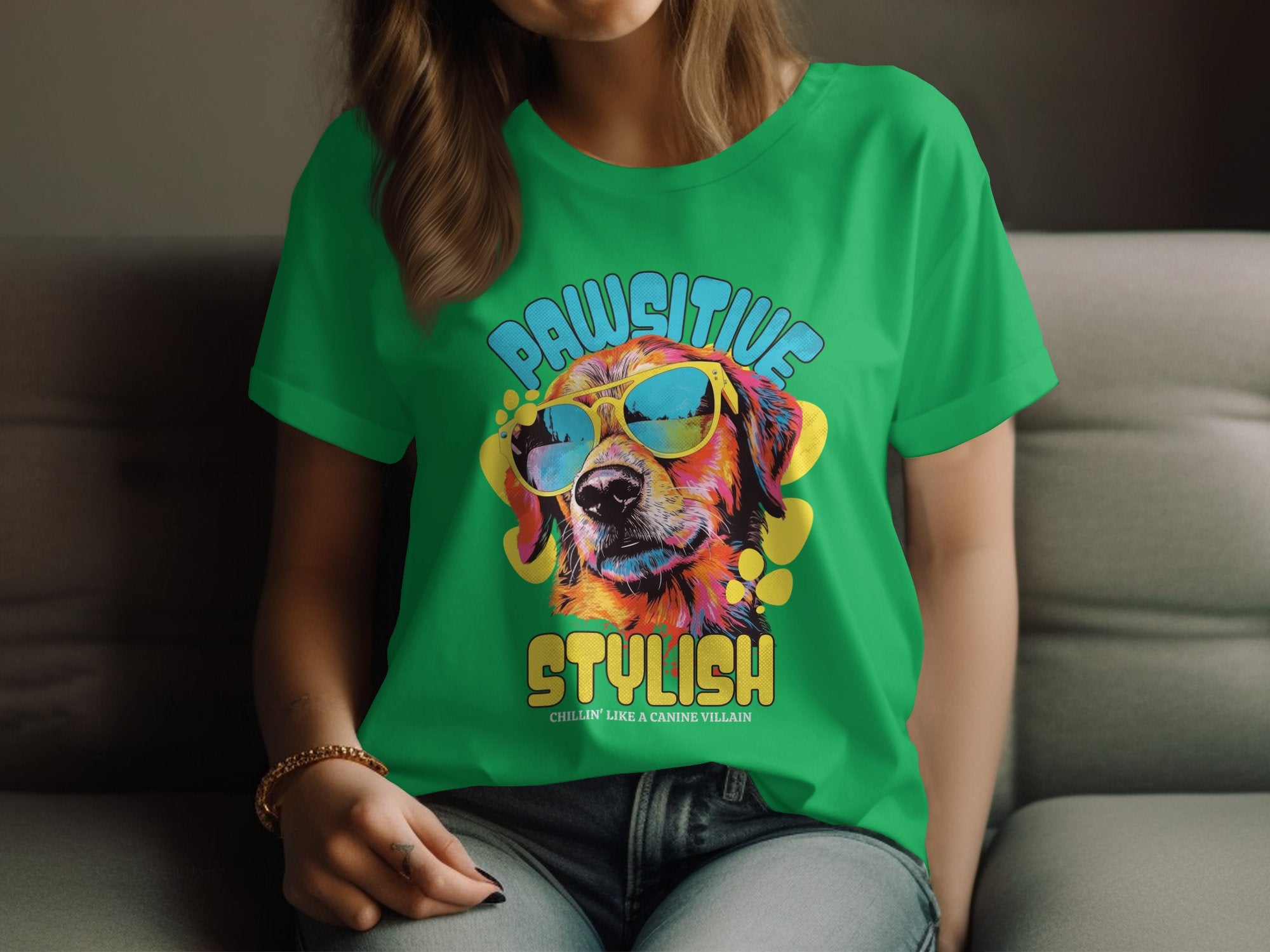 A person in a Garment Graphics green sunglasses t-shirt, featuring a colorful dog with shades illustration, sits with hands on their lap. The text says Pawsitive Stylish in womens sizing, with charming small text below.