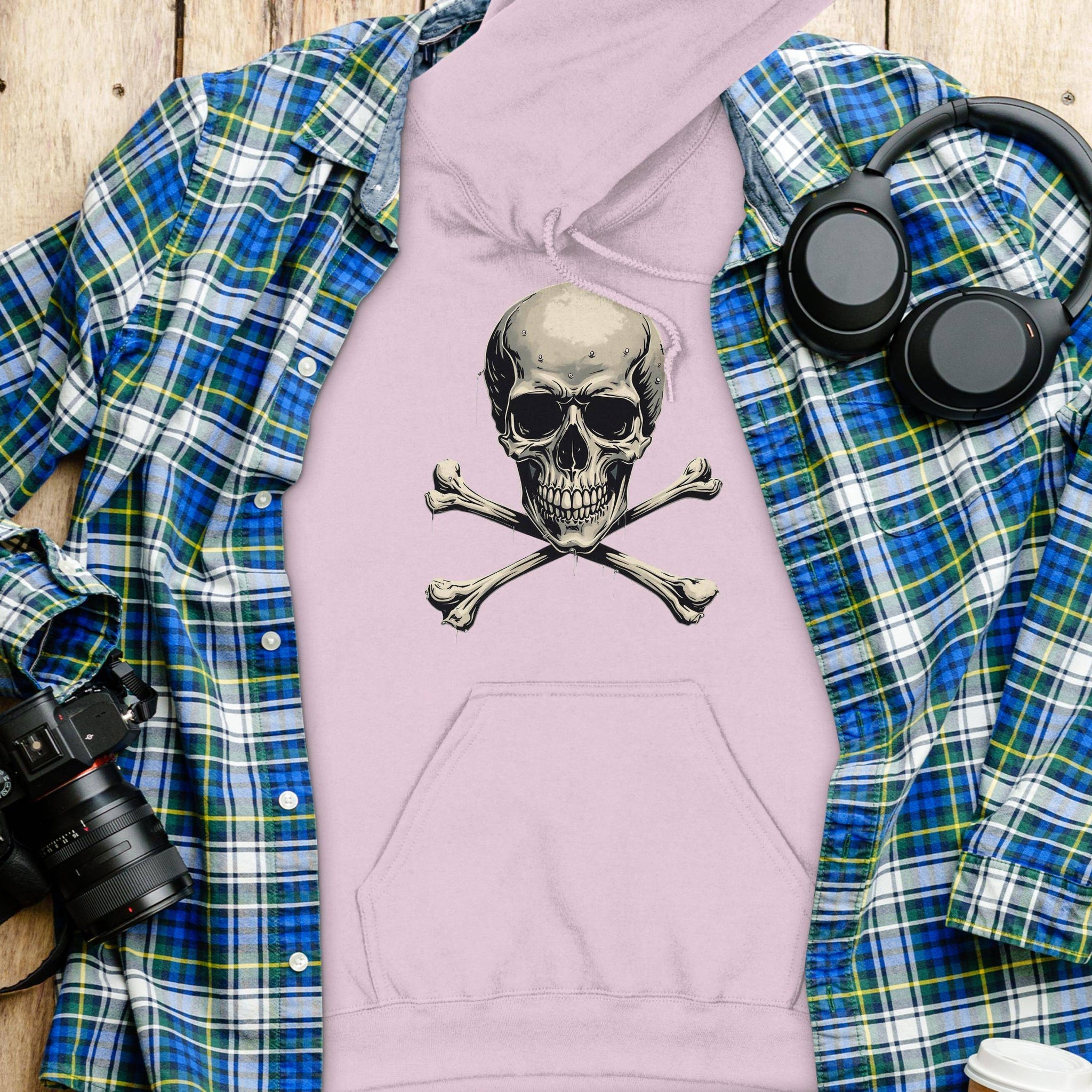 A fashion-forward flat lay showcases Garment Graphics blue and green plaid shirt layered over a pink hoodie with a skull and crossbones design. Nearby are black headphones, a camera, and a white water bottle on a wooden surface.