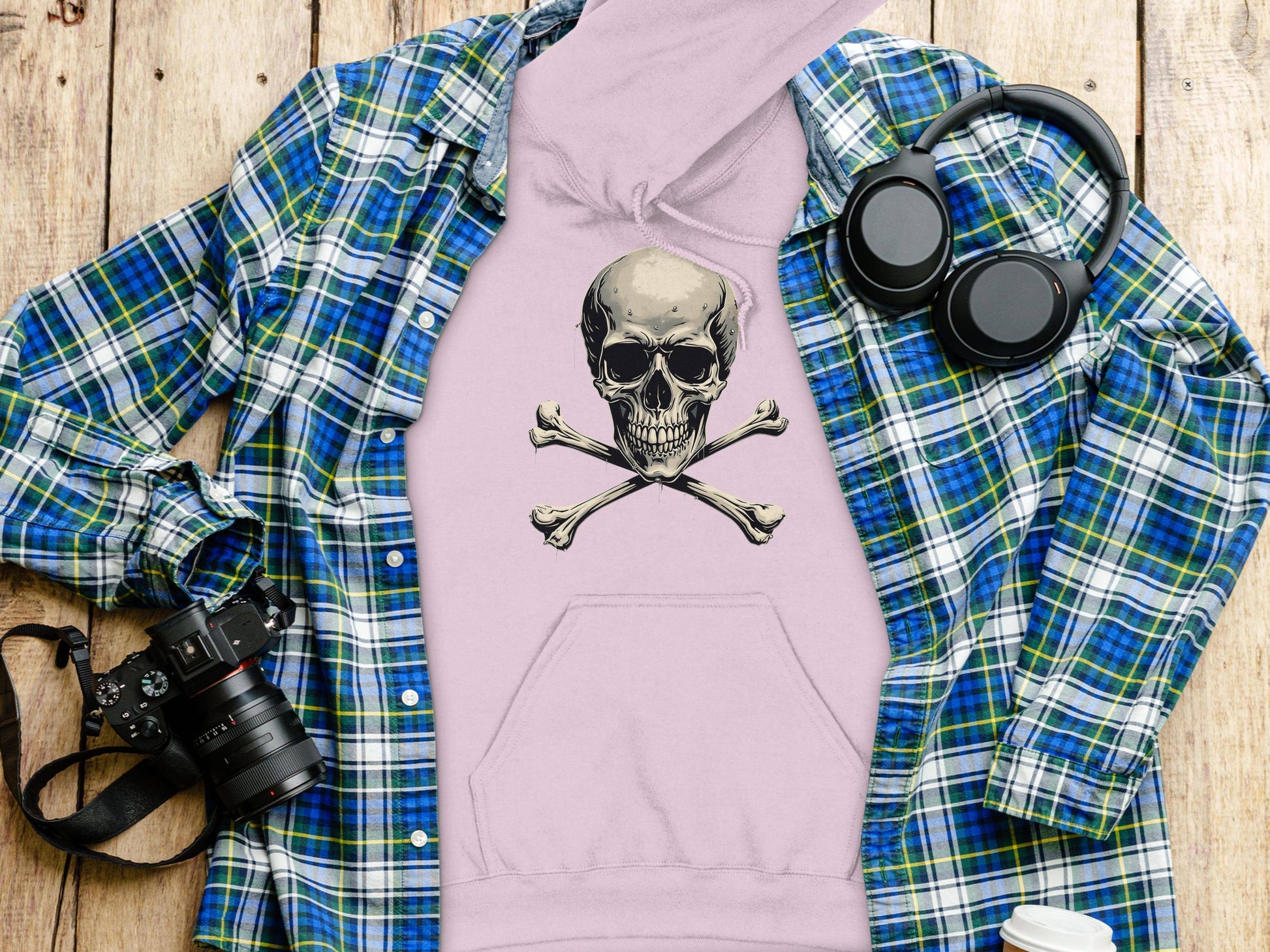 A fashion-forward flat lay showcases Garment Graphics blue and green plaid shirt layered over a pink hoodie with a skull and crossbones design. Nearby are black headphones, a camera, and a white water bottle on a wooden surface.