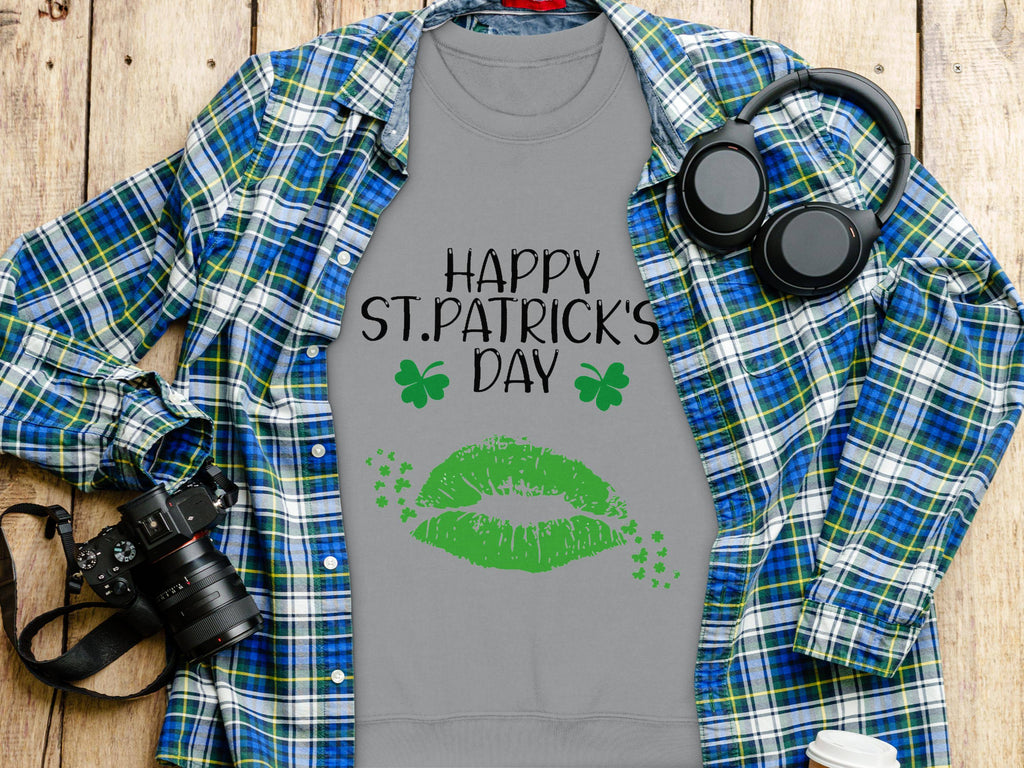 On a wooden surface, a gray Garment Graphics t-shirt boasts Happy St. Patricks Day in black, featuring a clover kiss with green lips and shamrocks. Its surrounded by a blue plaid shirt, headphones, a camera, notebook, and white cup—perfectly capturing the festive spirit.