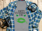 On a wooden surface, a gray Garment Graphics t-shirt boasts Happy St. Patricks Day in black, featuring a clover kiss with green lips and shamrocks. Its surrounded by a blue plaid shirt, headphones, a camera, notebook, and white cup—perfectly capturing the festive spirit.