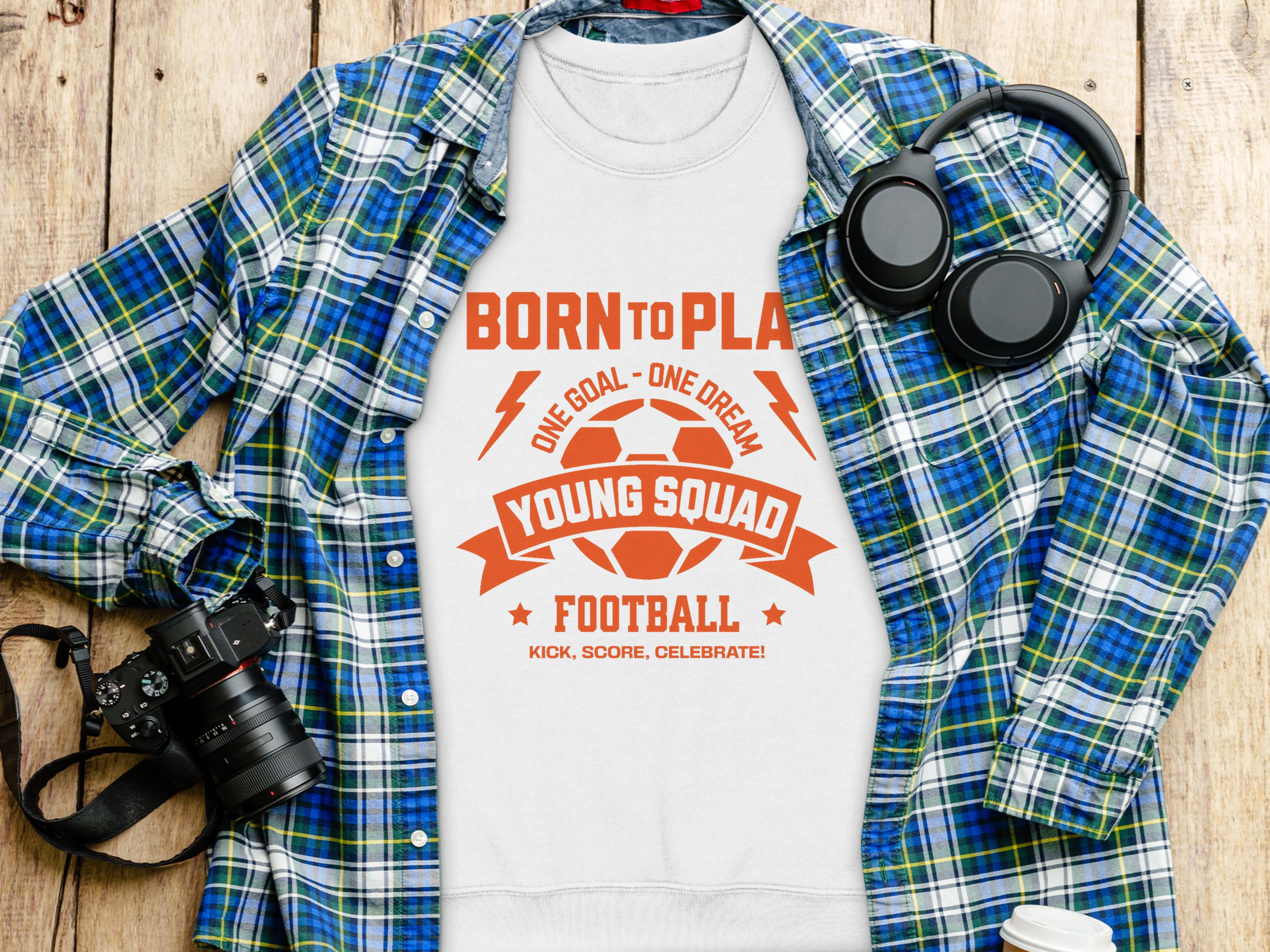 Garment Graphics presents a white unisex shirt featuring BORN TO PLAY in orange, football graphics, and ONE GOAL - ONE DREAM. Photographed with a blue plaid shirt, headphones, and camera on wood, its perfect for young football enthusiasts.