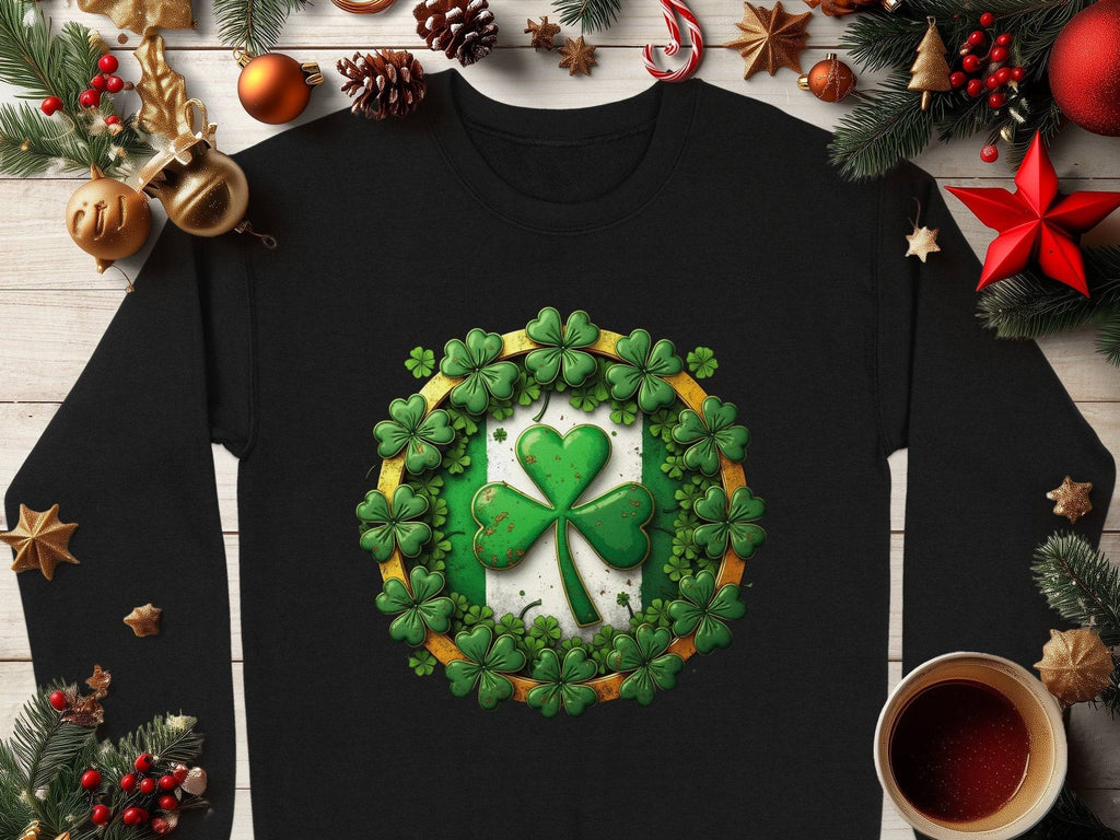 A Garment Graphics black long-sleeve shirt featuring a large green shamrock surrounded by smaller clovers lies on wood, adorned with Christmas ornaments, evergreen sprigs, and a nearby cup.