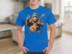 A person is wearing a blue Garment Graphics t-shirt, featuring a gorilla in sunglasses playing an electric guitar and making a peace sign with one hand. The background shows a blurred living room.