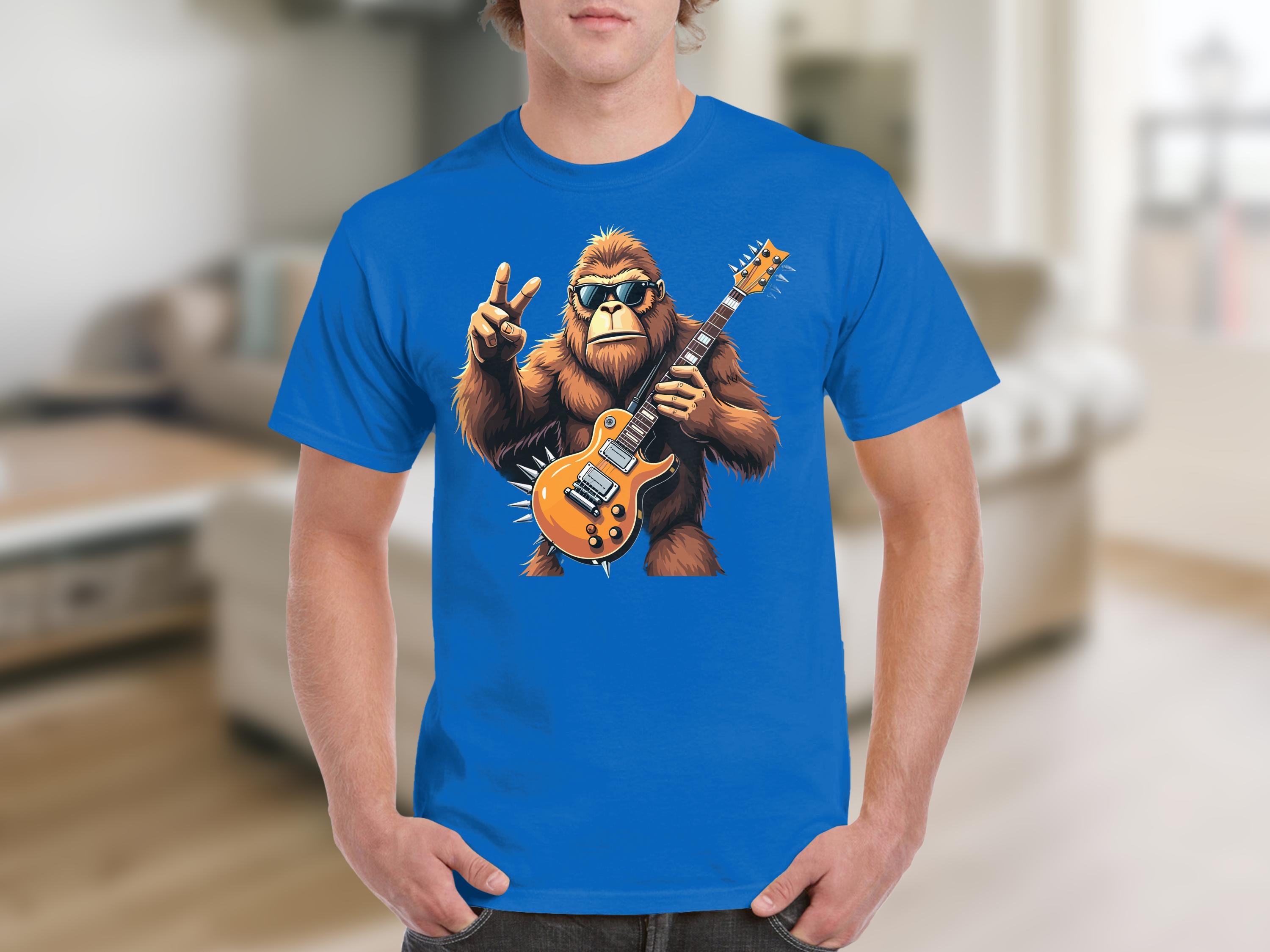 A person is wearing a blue Garment Graphics t-shirt, featuring a gorilla in sunglasses playing an electric guitar and making a peace sign with one hand. The background shows a blurred living room.