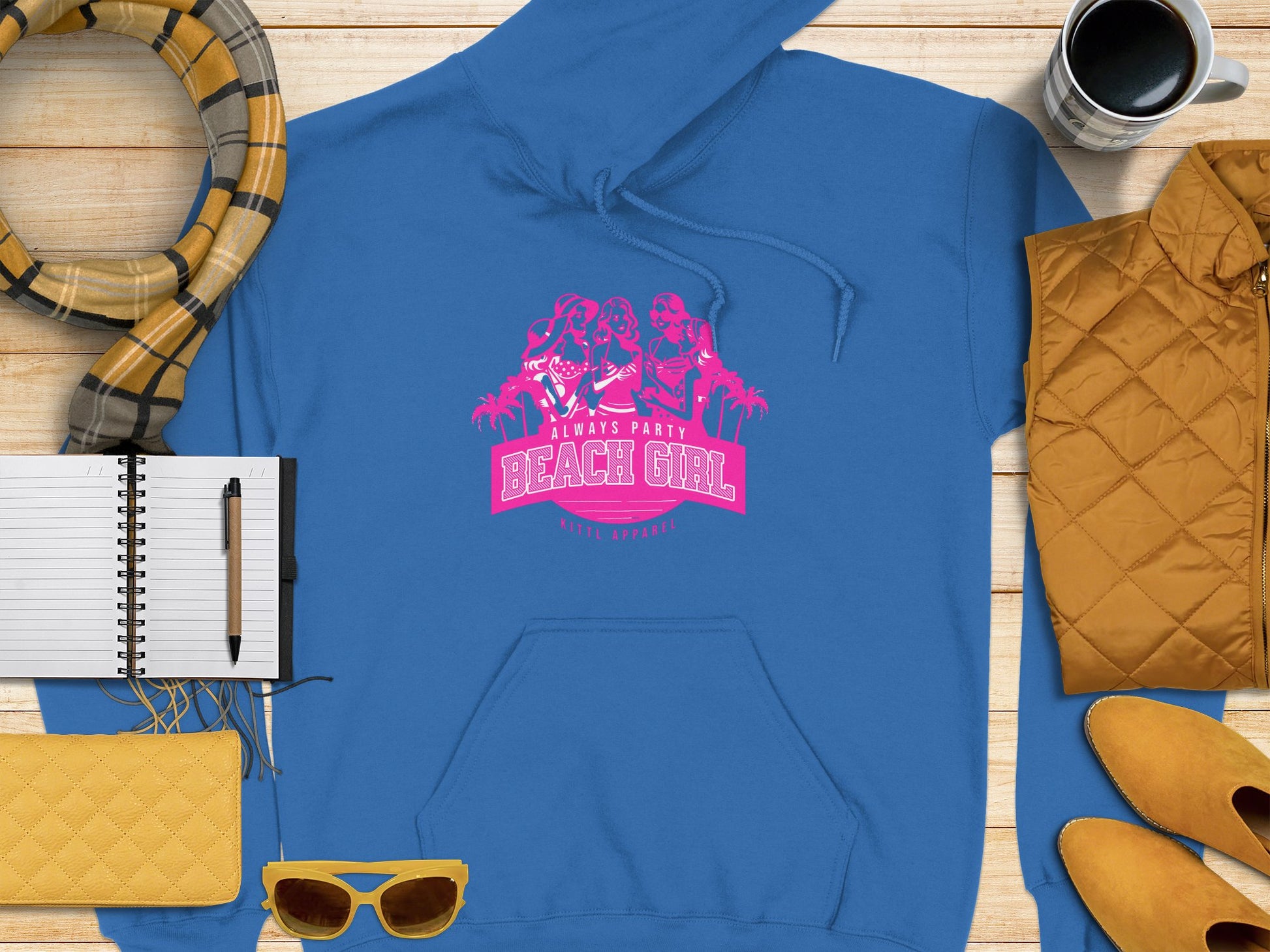 A vibrant blue Garment Graphics hoodie with Always Party Beach Girl in pink is stylishly displayed on wood, paired with yellow shoes, a matching purse, chic sunglasses, a patterned scarf, a notebook and pen, and a coffee cup for the perfect laid-back vibe.