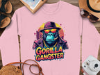 Garment Graphics urban pink sweatshirt showcases a gorilla graphic wearing a hat, sunglasses, and chain, with a cigar in its mouth. The text GORILLA GANGSTER appears below. Travel essentials like a straw hat, sunglasses, camera, and notepad complement the look.