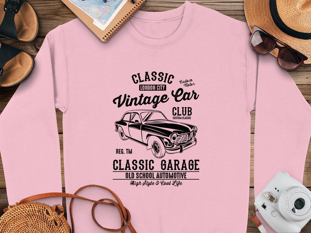 The Garment Graphics pink sweatshirt features a vintage car graphic with Classic Vintage Car Club text. Displayed with accessories like a notebook, sunglasses, straw hat, camera, and brown shoes on wood, its ideal for vintage car enthusiasts embracing classic London city automotive fashion.
