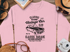 The Garment Graphics pink sweatshirt features a vintage car graphic with Classic Vintage Car Club text. Displayed with accessories like a notebook, sunglasses, straw hat, camera, and brown shoes on wood, its ideal for vintage car enthusiasts embracing classic London city automotive fashion.
