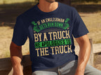Seated on a wooden bench outdoors, someone wears a navy blue Garment Graphics t-shirt with a clever design. The shirt humorously reads, If an Englishman gets run down by a truck, he apologizes to the truck, in green, white, and tan text.