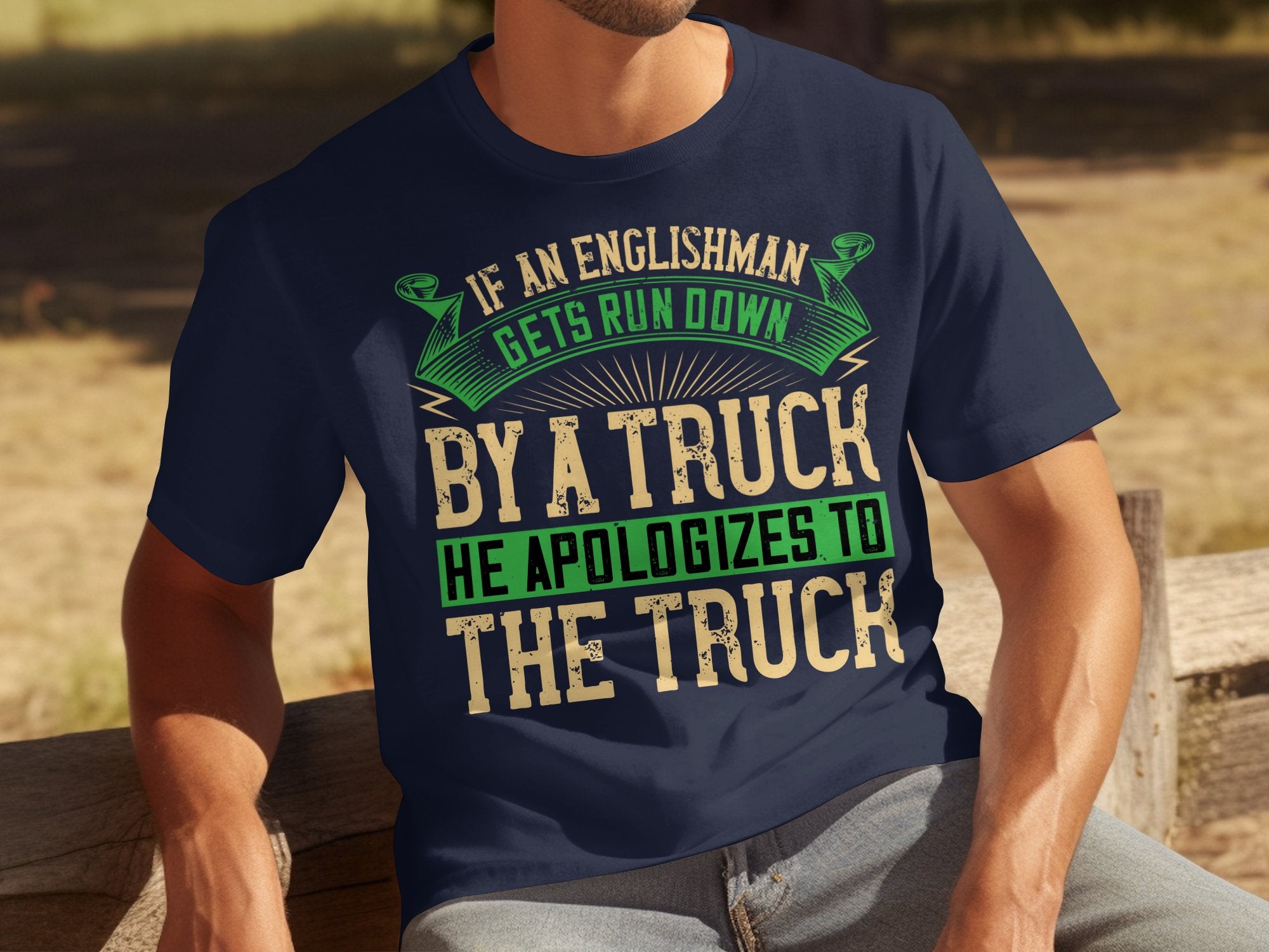 Seated on a wooden bench outdoors, someone wears a navy blue Garment Graphics t-shirt with a clever design. The shirt humorously reads, If an Englishman gets run down by a truck, he apologizes to the truck, in green, white, and tan text.