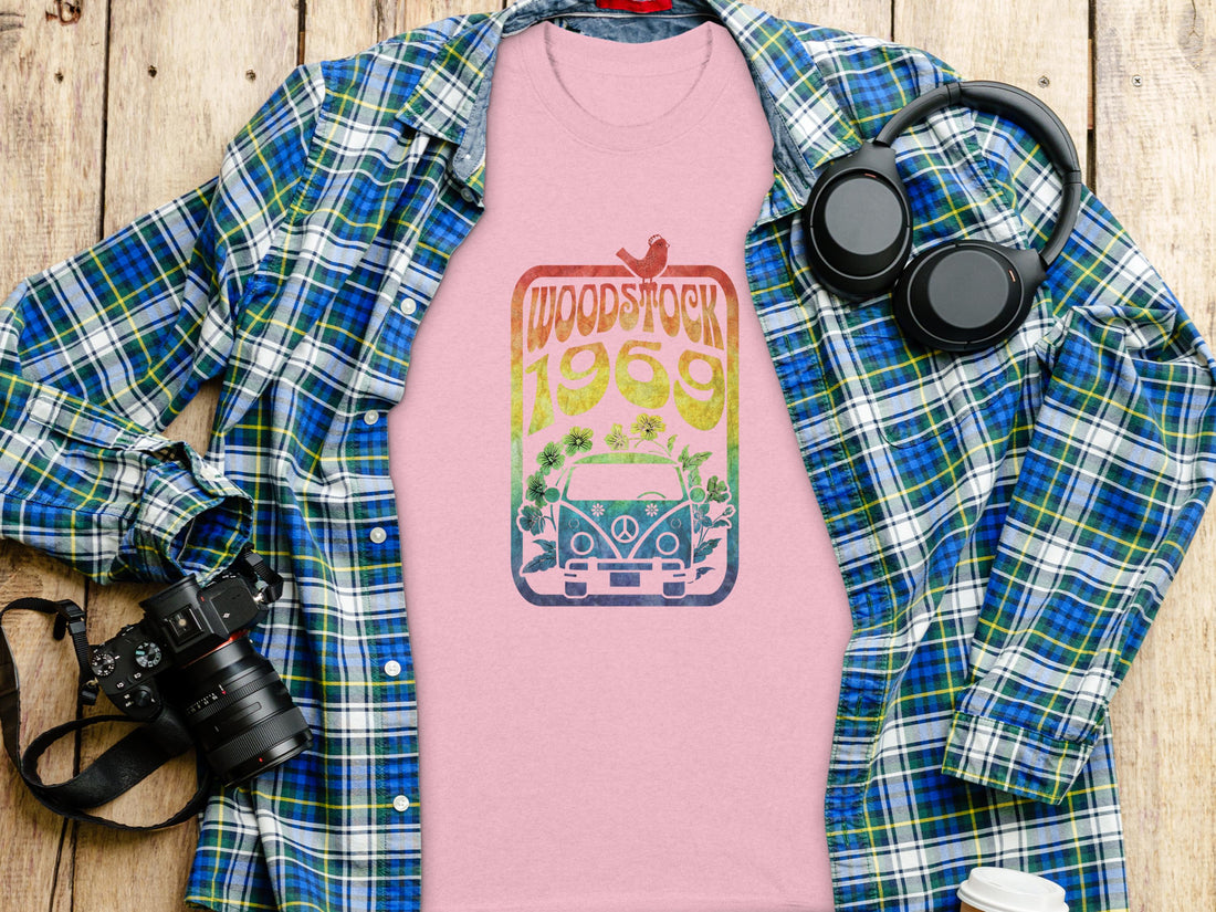 On a sandy beach, a music lover wears a Garment Graphics white T-shirt featuring Woodstock 1969 and a colorful retro van design. Tall grass sways gently in the breeze beneath the clear sky.