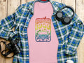 A pink Woodstock 1969 designer T-shirt by Garment Graphics rests on a blue plaid shirt on wood, paired with a camera, headphones, and white cup—ideal for music lovers. Every detail captures the free spirit of that retro van era.