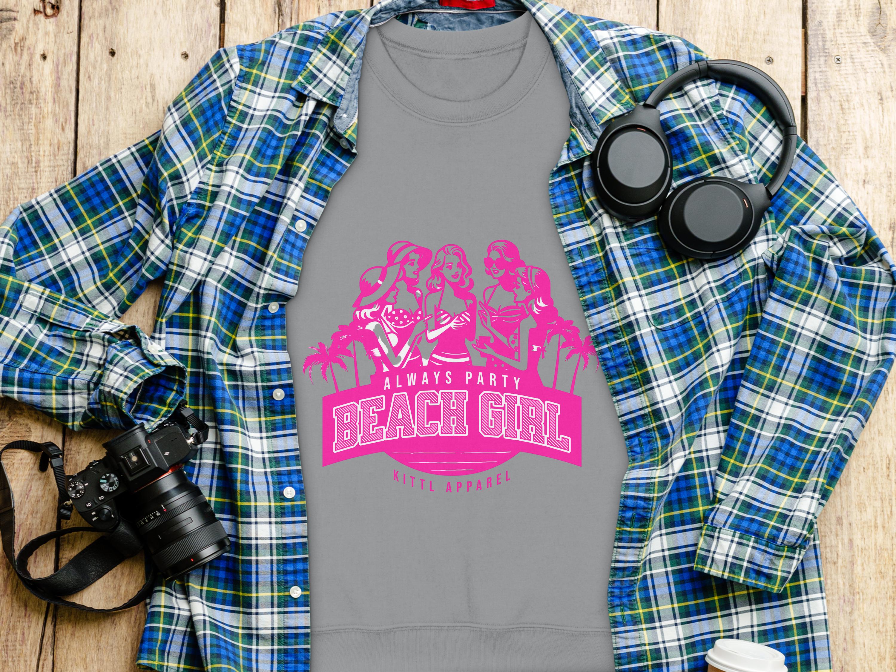 A gray Garment Graphics T-shirt featuring a pink Always Party Beach Girl design lies on a wooden surface, accompanied by a blue and green plaid shirt, black headphones, and a black camera.