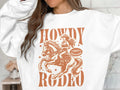 A person wears a white Garment Graphics sweatshirt featuring a graphic of a cowgirl on horseback. Bold Howdy Rodeo text and Not For Beginners in a badge design capture the cowboy spirit for true rodeo enthusiasts.