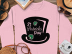 Garment Graphics presents a pink sweatshirt with a black top hat and green clovers graphic, featuring St Patricks Day text. It embodies festive spirit, displayed on wood and surrounded by accessories like a hat, sunglasses, shoes, notebook, and camera.
